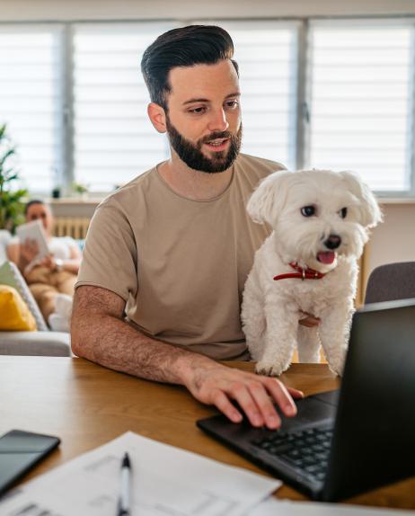 Man with dark beard with small white dog in his lap sits at home, working on a laptop with his wife in the background laying on a couch, reading.