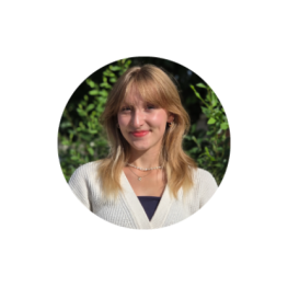 Communication student Communication Kenadie R. headshot - blonde woman with bangs and red lips smiling, wearing necklaces and a white cardigan, with greenery in the background.