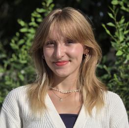 Kenadie R. headshot - blonde woman with bangs and red lips smiling, wearing necklaces and a white cardigan, with greenery in the background.