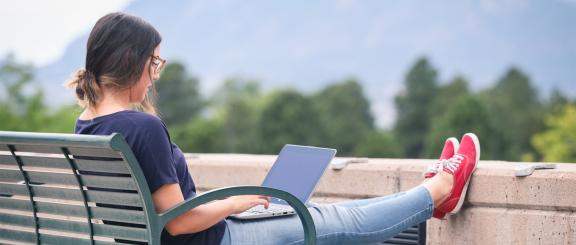 Woman looking at laptop sitting on bench with feet in red shoes up on cement ledge in front of her. Trees and Colorado Springs mountains in the background.