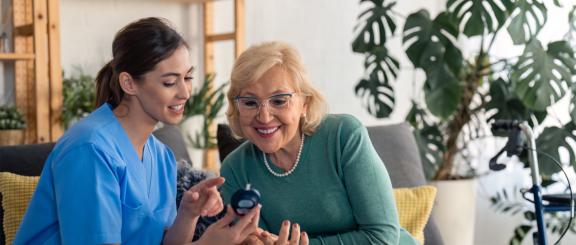 A young female health provider works with a middle-aged patient. The two subjects are looking at a blood sugar monitor and smiling.