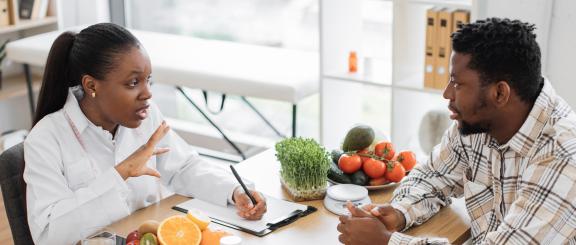 A female health provider in a lab coat takes notes as she works with a male patient. The two subjects are sitting at a table together and there are fruits and vegetables and a food scale set on the table.