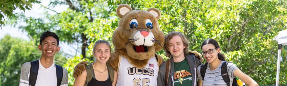 UCCS mascot, Clyde the Mountain Lion, wearing a white and gold basketball jersey, poses with arms around 4 young college students, smiling and wearing backpacks. Behind are bright green trees on a sunny day.