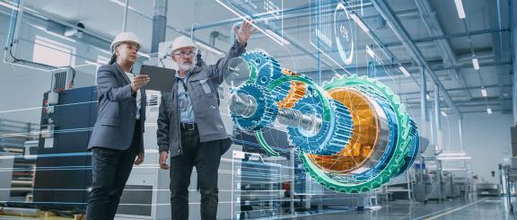 Man gestures his hand up towards a virtual, 3D display of a large, blue, green and orange mechanical cog equipment part. A woman in a navy blue blazer is next to him, holding a tablet, gazing on. Both wear white hard hats. Futuristic displays project all around around them.