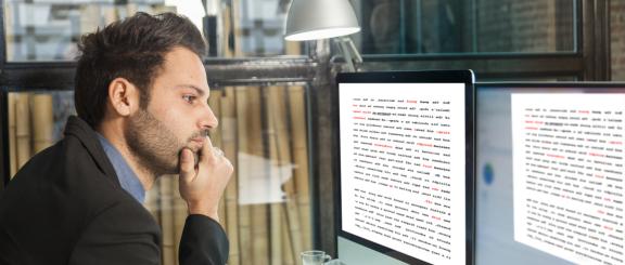 Man with fist on chin looks into 2 computer monitors, reading lots of rows of text on the screens.