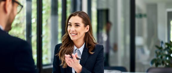 Brunette, long-haired woman wearing professional attire, smiles at man sitting across from her, as she motions her hand (holding a pen) towards him.