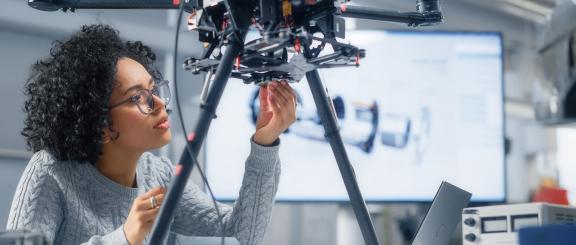 Curly, black-haired woman, wearing a grey sweater, looks at technical black machine on 2 legs, as she reaches under it to make adjustments.