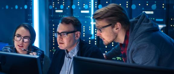 Woman and 2 men stare into 2 computer monitors with many gridded small round lights on a blue, glowing background, behind them.