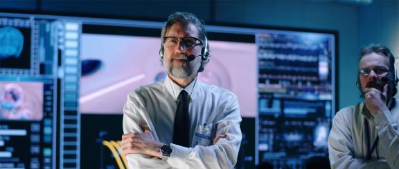 Man with greying beard and glasses, wearing headphones with an attached microphone, ID badge, crisp white shirt, tie and nice watch. His arms are crossed and he looks concerned with a control panel with many large screens behind him. Next to him is a man with his hand on his chin, with a furrowed brow.