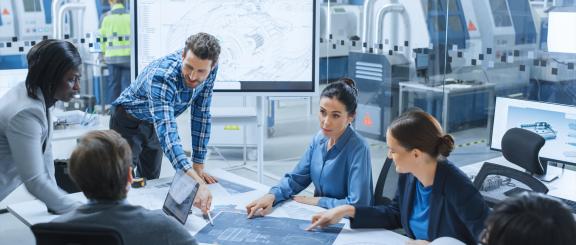 Man in plaid shirt leans over a table surrounded by a group of colleagues all looking at a large blueprint that he's pointing to.