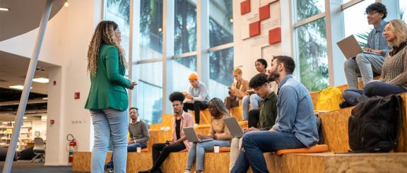 Woman in green blazer stands in front of group of seated young adult students looking up at her.