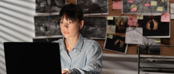 Woman sitting at desk working on laptop with many evidence pictures and sticky notes behind her on a bulletin board connected by string.