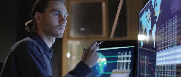 Man with hair tied back, looks concerned, points a pen in hand contemplatively at multiple computer monitors with graphics of continents and the globe.