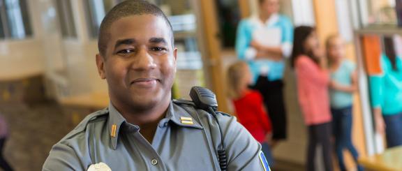 A man wearing a law enforcement uniform with a radio on his shoulder smiles at the camera while children gather in the background.