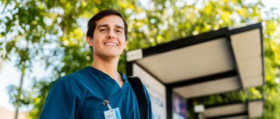 Nurse man with official id pass clipped to dark teal scrub shirt smiles in front of leafy green trees.