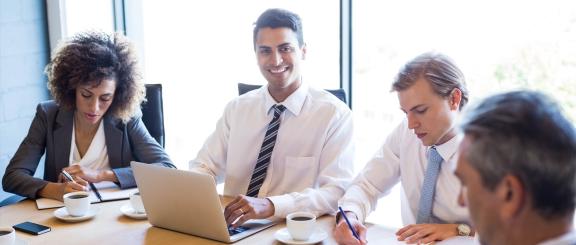 Man wearing a tie, sits at a table smiling, typing on a laptop with other well-dressed workers around him, busy writing on papers.
