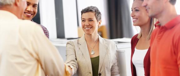 Woman wearing beige blazer and silver tear drop shaped necklace, with short greying hair, smiles as she shakes an older man's hand with other people surrounding them in an office setting.