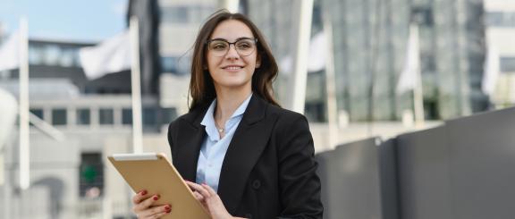 Woman smiling, wearing glasses and blazer with tablet in her hands. She stands in front of flags, with buildings in the background.