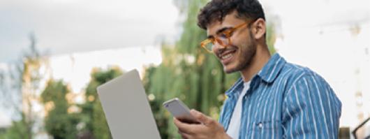 Smiling man outside with laptop and cell phone in hand.