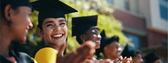 Row of graduates smiling and clapping, wearing black regalia and square-top hats with tassels.