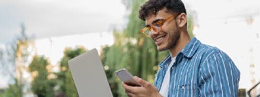 Smiling man outside with laptop and cell phone in hand.