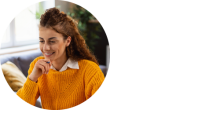 Woman in orange sweater looks into computer smiling, book on the desk in front of her, with her hand on her chin.