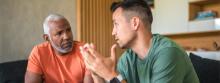 2 men sit together looking at each other seriously. The white haired older man looks at the younger man with understanding and concern.