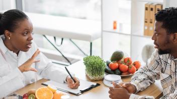 Woman in white with measuring tape hanging around her neck, writing on clipboard, explaining nutrition to man sitting across from her, with vegetables on their table.