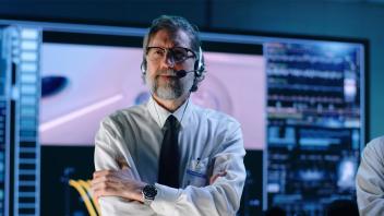 Man with greying beard and glasses, wearing headphones with an attached microphone, ID badge, crisp white shirt, tie and nice watch. His arms are crossed and he looks concerned with a control panel with many large screens behind him. Next to him is a man with his hand on his chin, with a furrowed brow.