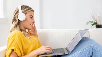 Smiling blonde woman wearing white headphones and gold/yellow shirt, sits on her couch with a laptop in her lap, in a bright home.