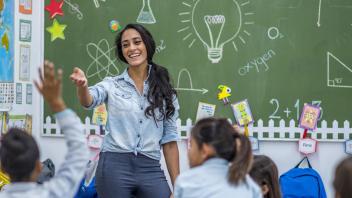Smiling teacher woman in denim shirt with brunette hair gestures her arm towards an elementary school student boy, raising his hand to be called on. Behind her is a green chalkboard with many symbols drawn in white - such as a light bulb, atom, triangle, graph, science beaker, math equation, ect. and a world map on the wall. Many other young student kids sit in their desks in front of their teacher.