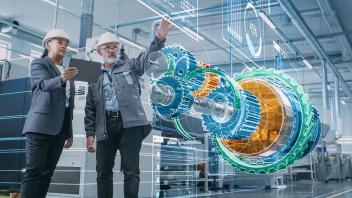 Man gestures his hand up towards a virtual, 3D display of a large, blue, green and orange mechanical cog equipment part. A woman in a navy blue blazer is next to him, holding a tablet, gazing on. Both wear white hard hats. Futuristic displays project all around around them.