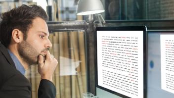 Man with fist on chin looks into 2 computer monitors, reading lots of rows of text on the screens.