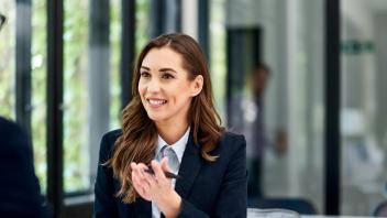 Brunette, long-haired woman wearing professional attire, smiles at man sitting across from her, as she motions her hand (holding a pen) towards him.