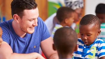 Young teacher man smiles at many children around him.
