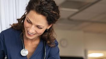 Nurse woman with brunette hair with stethoscope around neck looks down smiling at unseen patient.