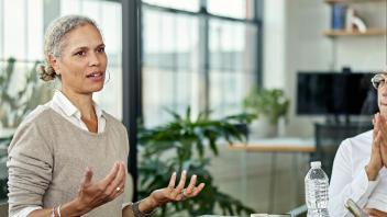Woman with greying hair and smart attire looks concerned, with both palms raised slackly in front of her. They sit in an office, co-worker nearby, looking on with round glasses and hands in a prayer gesture.