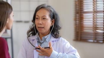 Asian nurse woman with greying shoulder length hair wearing a lab coat and stethoscope in ears with her hand over her lungs, sitting with a brunette young patient in dark red shirt.
