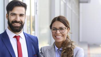 Outside of a glass-lined building, a hansom man with thick beard, wearing a blue suit with red tie, smiles next to a happy, brunette, woman next to him, wearing a grey blazer.