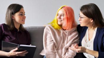 Social worker woman sitting on couch, looking over at concerned mother and smiling daughter (with brightly colored hair) next to her.