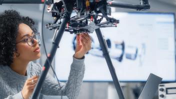 Curly, black-haired woman, wearing a grey sweater, looks at technical black machine on 2 legs, as she reaches under it to make adjustments.