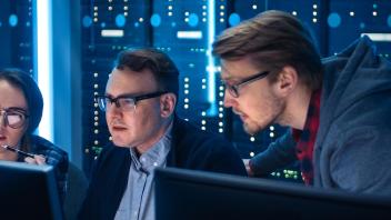 Woman and 2 men stare into 2 computer monitors with many gridded small round lights on a blue, glowing background, behind them.