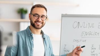 Man smiles at phone camera on tripod in front of him. He points to a dry erase board with "Online Education English Lesson" writing.