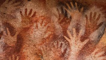Many handprints outlined with dark orange and red paint on cave wall.