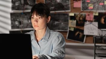 Woman sitting at desk working on laptop with many evidence pictures and sticky notes behind her on a bulletin board connected by string.