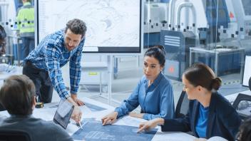 Man in plaid shirt leans over a table surrounded by a group of colleagues all looking at a large blueprint that he's pointing to.