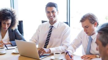 Man wearing a tie, sits at a table smiling, typing on a laptop with other well-dressed workers around him, busy writing on papers.