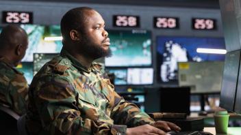 Man wearing camo sits in control room looking into many computer monitors.