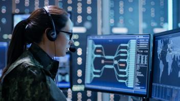 Woman wearing camo, glasses and headphones, looks into multiple computer monitors showing complex maps.