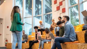 Woman in green blazer stands in front of group of seated young adult students looking up at her.